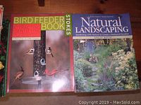 Two books shown side by side on a wooden floor, Birdfeeder Book and Natural Landscaping, covers visible with birds and garden imagery.
