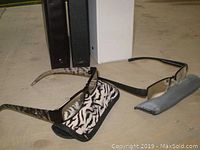 Two pairs of reading glasses with their respective soft cases placed on a flat surface, with three binders positioned behind them.
