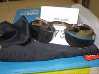 Silver steampunk glasses with closed flip-up lenses, resting on a black cloth with a small black pouch beside them.