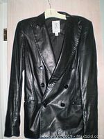 Front view of the black leather jacket showing double-breasted buttons and notched lapel collar, hanging on a white hanger against a door.