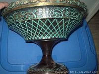 Side view of a heavy metal bowl on pedestal showing green patina, floral border, lattice pattern, and ornamental rim with some chipping.