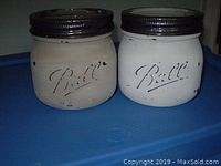 Two small painted Ball mason jars side by side showing front embossed logo, white paint with black screw lids.