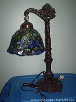 Full view of old style table lamp with colorful stained glass style shade, ornate dark metal arm and base, on blue surface.