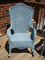 Blue wicker rocking chair on wooden deck under sunlight, showing the seat, armrests, and top of the backrest with woven diamond pattern.