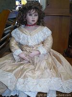 Full view of the seated vintage ceramic doll wearing a cream-colored dress with lace detailing and red bead necklace.