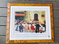 Framed print showing multiple people dining outside a restaurant labeled 'Restaurant' and a hotel labeled 'Hotel'. Colorful lively scene with greenery, children, adults, and a dog visible.