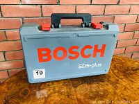 Closed gray Bosch branded plastic carrying case with red latches sitting on wooden table in front of brick wall.
