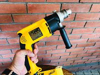 Photo showing yellow and black DeWalt DW511 hammer drill held in hand against brick wall, includes side handle and metal chuck
