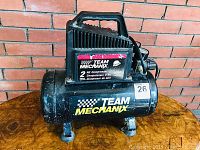 Front view of the Team Mechanix air compressor placed on a wooden surface against a brick wall, showing the full item with carrying handle and tank.