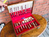 Opened wooden chest displaying silver plated knives, forks, spoons, and teaspoons in red and white lined slots.