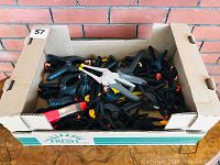 Wide view of the cardboard box filled with assorted clamps including black spring clamps and a metal staple gun style clamp