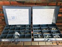 Two open steel fastener bins showing organized compartments filled with metal screws, bolts, anchors, and labels inside lids.