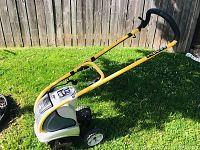 Side view of Ryobi electric garden tiller on grass showing yellow handlebar and rear wheels.