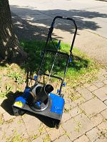 Front and side view showing the Snowjoe snow blower with chute and handlebar visible, placed on paved surface under sunlight.