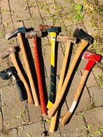 Eight axes and hatchets arranged on a paved surface showing varying handle types and head conditions.