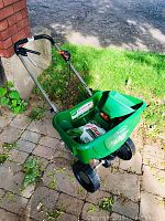 Green Scotts Turf Builder broadcast spreader loaded with a bag of fertilizer and a handheld spreader inside it sitting outside on a brick patio and grass.
