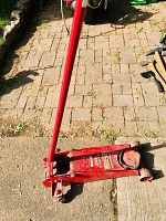 Angle view showing floor jack with long red handle propped vertically, outdoor setting on stone tile.
