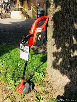 Trimmer edger leaning against a tree outdoors, showing the full tool with red and black color scheme.