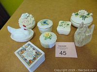 Seven assorted trinket boxes including ceramic swan, chicken, floral designs and clear glass animal-shaped box arranged on brown surface