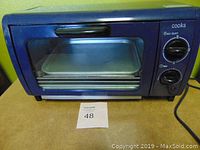 Front view of blue Cooks toaster oven with heat select and timer control dials and visible metal baking trays inside.
