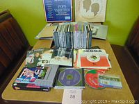 Photo showing a collection of vinyl records, a stack of CDs, DVDs, and cassette tapes spread on a table with some visible titles and sleeves.