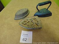 Photo showing two vintage sad irons and an ornate metal stand on a table, depicting the entire lot.