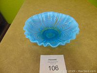 Top-front angled view of the blue opalescent ruffled bowl placed on a tan surface with a MaxSold label in front, showing the ruffled edge and swirl pattern.