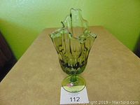 Green glass handkerchief-shaped vase on beige surface against green wall. Shows wavy flared rim, vertical ridges, round flat base with Fenton label.