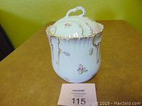Porcelain biscuit jar with lid featuring floral designs and gold rimning on a table, MaxSold lot card present.