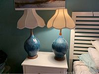 Two 32 inch lamps with blue and green earth pattern bases and cream clover-shaped shades side by side on a white nightstand under a wall painting.