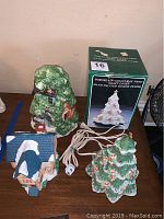 Three holiday ceramic decorations: a green lighted Christmas tree with cord, a boxed white porcelain Christmas tree, and a ceramic holiday house with snow accents.