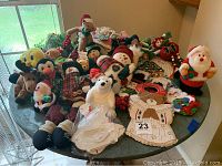 Photo showing a collection of assorted Christmas plush decorations arranged on a round table near a window. Includes multiple Santa Claus figures, reindeer, bears, snowmen, and other holiday plush items along with some fabric decorations on the table.