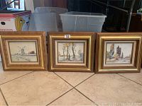 Three framed watercolor paintings on tile floor, showing countryside and nature scenes with wooden gold-toned frames