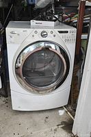 Front view of white Whirlpool dryer with chrome door trim, showing general wear and surrounding garage items.