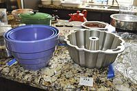 Three nested purple plastic mixing bowls beside metal bundt pan on speckled countertop with other kitchen items.