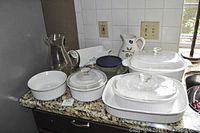 Image shows a variety of white Corningware dishes including a loaf dish and round dishes with glass lids, assorted pitchers, and a glass tray with plastic lid placed on a granite countertop.