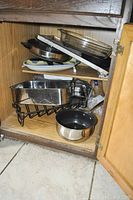 Metal pots, pans, baking trays stacked inside a wooden kitchen cabinet under the sink area.