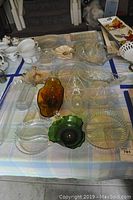Table covered with checkered cloth with multiple glass bowls and trays in clear, amber, green and pink glass. Various shapes and serving uses.