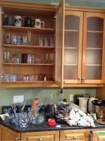 Wide view of kitchen countertop and open cabinets showing various glasses, mugs, and barware items scattered on counter and shelves.