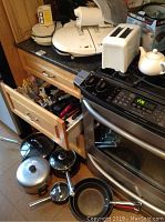 Kitchen counter and stove area with George Foreman grills, toaster, frying pans, pots, and open drawers showing kitchen utensils and knives.