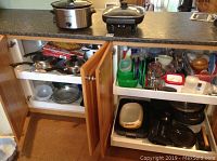 Wide view of kitchen cabinet open showing various pots, pans, glassware, bowls, and kitchen utensils inside and on shelves