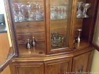 Wooden cabinet with multiple types of clear stemware glasses and silver plated items arranged inside, showing general lot contents