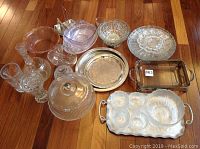 Overview of lot showing multiple clear and frosted glass dishes, cake plate with lid, trays, and pitchers arranged on wooden floor.