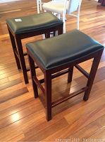Pair of two bar stools shown from angle with black leather cushions and dark wooden legs on wooden floor