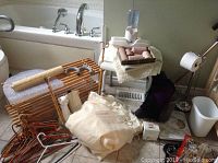 Photo showing items including bath soap dispenser, shoe rack, hangers, toiletries, heating pad, scale on tile floor near bathtub, includes toilet paper holder and white waste bin.