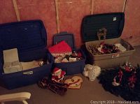 Three large plastic bins with lids off showing various Christmas decorations including wreaths, Santa figurine, and ornaments on concrete floor in unfinished room.