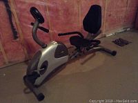 Wide side view of the FreeSpirit 803 recumbent stationary bike showing the silver main body, black seat and backrest, pedals, and part of the handlebars.