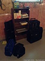 Photo of wooden bookshelf with assorted books, silver metal tray shelf, two black luggage bags beside it, and two vertical black CD racks leaning against the wall.