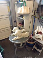 Photo showing white wooden shelving unit with multiple shelves, resting against a wall, with lion garden statues and patio table in front.