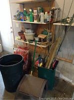 View of shelf with various garden chemical sprays, multiple plant pots stacked, bags of mulch, garbage bin, and garden tools leaning against the shelf